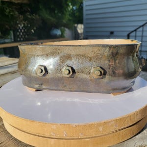 May include: A handmade, rectangular ceramic planter with a metallic bronze glaze. The planter features three decorative bolt accents on the front and small feet. The interior has a lighter, beige colour.