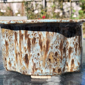 Puede incluir: Una jardinera rectangular de cerámica con un esmalte texturizado único. La jardinera presenta una base azul claro con detalles marrones y dorados, creando un efecto veteado. El borde es de color marrón oscuro. Diseñada para uso interior o exterior.