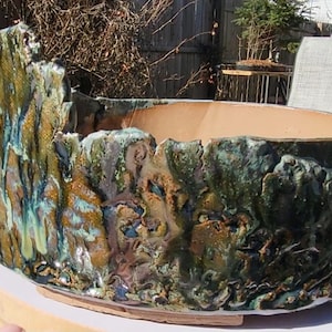 Puede incluir: Una maceta de cerámica grande, de forma ovalada, con un exterior texturizado y multicolor. La maceta presenta un diseño rugoso, similar a la corteza, con tonos verdes, marrones y azules. El interior es de color marrón claro. La maceta está sobre una base redonda y clara.