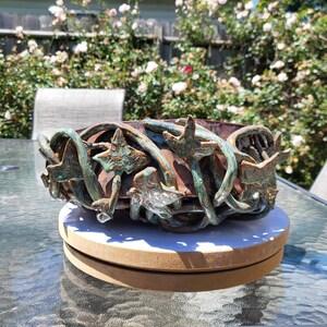 May include: A round ceramic bowl with a dark brown base, featuring an intricate exterior design of sculpted, three-dimensional green ivy vines and leaves that wrap around the entire circumference of the vessel, creating a textured, organic appearance.