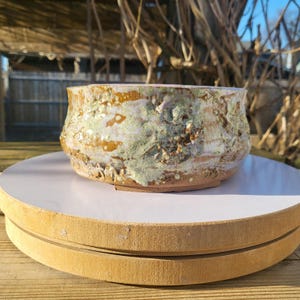 May include: A handcrafted ceramic bowl with a textured surface. The bowl features a blend of colors, including brown, white, and green, creating a unique, organic appearance. The bowl is sitting on a white surface.