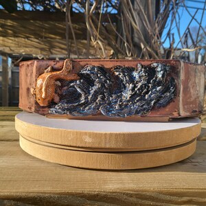 Puede incluir: Una maceta rectangular de cerámica para bonsái con acabado marrón rojizo. La parte frontal presenta un relieve tridimensional de un pez koi de color naranja dorado nadando entre olas oscuras, texturizadas y de color azul negruzco brillante.