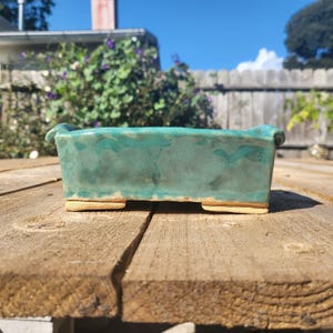 Op de afbeelding: Een rechthoekige, turquoise keramische plantenbak met een getextureerd oppervlak. De plantenbak heeft een lichtbruine basis en kleine voetjes. De achtergrond omvat een houten oppervlak, groen en een heldere blauwe lucht.
