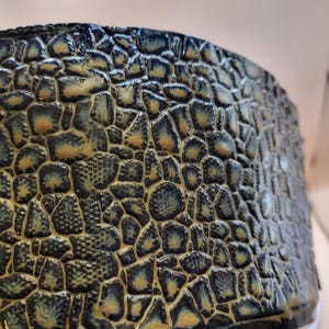 May include: Close-up of a ceramic vessel with a textured, mosaic-like pattern. The design features a dark blue base with irregular, raised shapes in shades of tan and blue, creating a unique, tactile surface. The vessel's rim is visible.