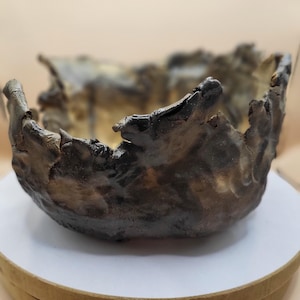May include: A handcrafted, organic-shaped bowl with a rough, textured surface. The bowl features a dark brown exterior with hints of gold and a jagged rim. The interior appears to have a golden hue. The bowl is likely made of ceramic or a similar material.