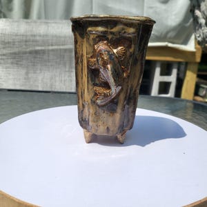 May include: A cylindrical ceramic planter with a glossy, mottled brown and tan glaze. The exterior features a raised, sculpted koi fish relief in the center. The vessel is supported by three small, pointed feet at the base, providing stability.