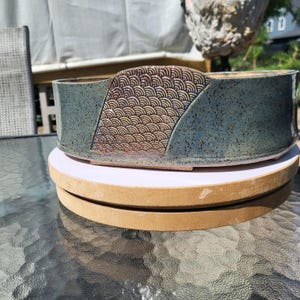 May include: A low, wide ceramic bonsai pot with a speckled dark teal glaze. The exterior features a prominent, raised triangular panel decorated with a repeating, incised fan-shaped wave pattern. The pot has a smooth, flat rim and a circular base.