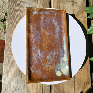 May include: A rectangular ceramic tray with a rustic, mottled brown glaze featuring subtle, light green and blueish-white highlights across the surface. The tray has slightly raised edges and a textured, uneven finish with visible speckling.