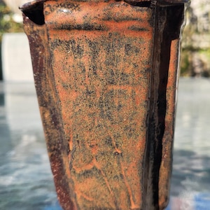 Könnte beinhalten: Eine handgefertigte Keramikvase mit einer einzigartigen, strukturierten Oberfläche. Die Vase hat eine quadratische Form mit leicht ausgestelltem Oberteil und eine reichhaltige Farbpalette aus Braun, Orange und Schwarz. Die Vase wird von kleinen, rechteckigen Füßen getragen.