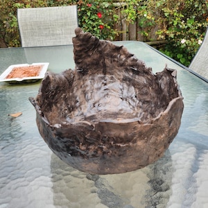 May include: A handcrafted, dark brown ceramic bowl with a textured, organic design. The bowl has a rough, uneven rim and a glossy finish. The bowl is sitting on a glass table outdoors.