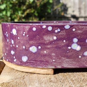 May include: A handmade ceramic bowl with a deep purple glaze and speckled white and light green accents. The bowl has a rounded rectangular shape and sits on a natural clay base. The speckled design gives the bowl a unique, artistic look.