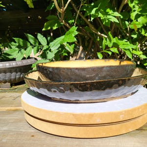May include: Two stacked oval ceramic bowls with a dark, glossy, mottled brown and black exterior glaze and a smooth, matte, light yellow interior. The bowls feature an irregular, hand-formed rim and a textured, wavy surface along the outer sides.
