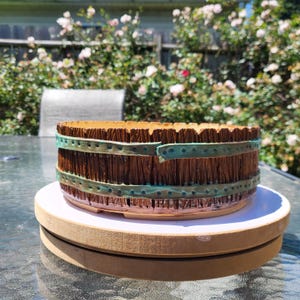 May include: A round, low-profile planter with a textured brown exterior resembling vertical wooden planks. Two horizontal, teal-colored bands with a repeating dot pattern wrap around the circumference of the planter, mimicking the appearance of metal straps.