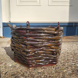 Puede incluir: Una cesta de cerámica cuadrada hecha a mano con un diseño tejido. La cesta está esmaltada en tonos marrones y burdeos, con un acabado brillante. La cesta es adecuada para la decoración del hogar.