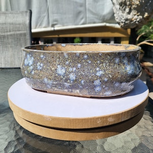 May include: An oval ceramic bonsai pot with a speckled glaze featuring shades of gray, blue, and yellow. The exterior surface has a textured, mottled appearance with a smooth, curved rim. The interior of the pot is a light, unglazed tan color.