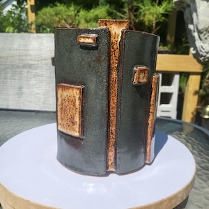 May include: A cylindrical ceramic vase featuring a dark, glossy charcoal-grey glaze contrasted by textured, speckled tan rectangular accents. The surface displays vertical seams and raised geometric patches with a rough, earthy finish.