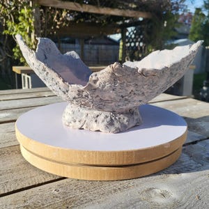May include: A handcrafted, textured bowl with a unique, organic shape. The bowl is primarily light gray with subtle pink undertones, and sits on a white and wood-toned rotating display stand. The bowl's rim has an uneven, sculptural edge.