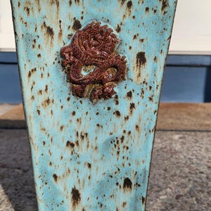 May include: A turquoise ceramic vase with a brown dragon relief. The vase has a speckled pattern and a rectangular shape that tapers towards the base. The dragon is detailed and adds a decorative element.