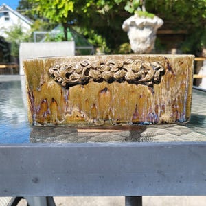 May include: A rectangular ceramic planter with a decorative floral relief. The planter has a glazed finish with shades of gold, brown, and purple. The planter is designed for indoor or outdoor use.