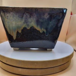 Puede incluir: Una jardinera cuadrada de cerámica con un esmalte azul oscuro, verde azulado y dorado. La jardinera tiene una forma ligeramente acampanada y se apoya en cuatro pequeños pies cuadrados. El esmalte tiene un efecto de goteo, creando una apariencia texturizada única.