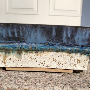 May include: Rectangular ceramic planter with a speckled off-white base and a dark blue-glazed top. The planter has a turquoise rim and sits on a wooden base, suitable for indoor or outdoor use.