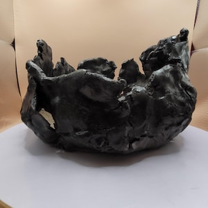 May include: A dark, textured, bowl-shaped sculpture with an organic, rough appearance. The black object has an uneven rim and a textured surface, suggesting a handcrafted or natural aesthetic. The bowl sits on a white surface.