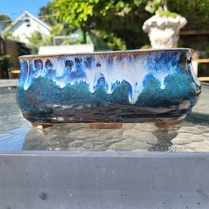 Puede incluir: Una jardinera rectangular de cerámica con acabado brillante. La jardinera presenta un degradado de colores, que va del azul oscuro en la base al turquesa y blanco en el borde. El borde tiene un acento marrón. La jardinera se apoya en pequeños pies de madera.