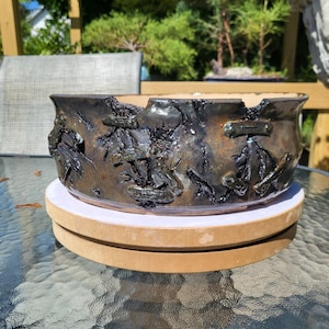 Puede incluir: Una maceta de cerámica para bonsái, baja y ancha, con un esmalte metálico en tonos bronce oscuro y negro. La superficie exterior presenta un acabado rugoso y texturizado con elementos decorativos en relieve en forma de barra.