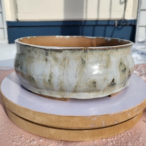 May include: A handmade ceramic bowl with a wide, shallow design. The bowl features a light, speckled glaze with brown and gray accents. The interior is a warm, earthy brown. The bowl sits on a wooden base, perfect for serving or display.