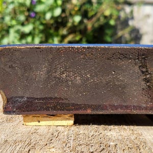 May include: A rectangular, dark brown ceramic planter with a textured surface. The planter is supported by a small, light-colored block. The background is blurred, showing greenery.