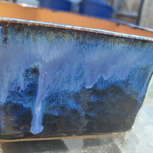 May include: Rectangular ceramic planter with a glossy, dripped glaze in shades of blue and black. The interior is a warm, earthy tone. The planter is suitable for indoor or outdoor use.