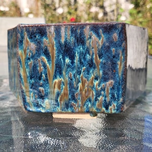 May include: A square ceramic planter with a glossy finish. The planter features a blend of blue, teal, and brown colors, creating a unique, textured appearance. The planter is designed with a modern, geometric shape, suitable for various plants.