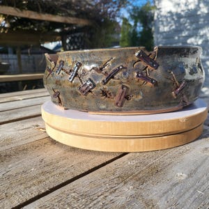 May include: A handmade ceramic bowl with a unique design. The bowl is a mix of brown and blue-green hues, with dark brown rectangular accents. The bowl sits on a light-colored wooden base, showcasing its handcrafted nature.