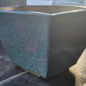 Puede incluir: Una maceta cuadrada de cerámica con un exterior moteado turquesa y gris. La maceta tiene una superficie ligeramente texturizada y un interior gris oscuro. La maceta está sobre una superficie oscura.