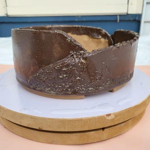 Puede incluir: Una maceta de cerámica marrón hecha a mano con un exterior texturizado y rugoso. La maceta tiene un diseño asimétrico único con una sección recortada en el borde. El interior es de color marrón claro. Ideal para suculentas o plantas pequeñas.