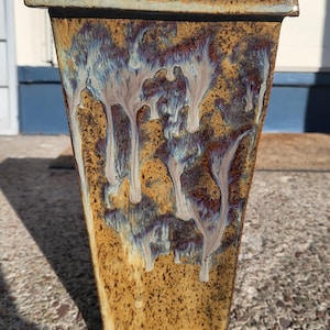 May include: A tall, rectangular ceramic planter with a unique glaze. The planter features a blend of brown, gold, and blue hues with a dripped pattern. The top rim is a darker brown. Suitable for indoor or outdoor use. Measurements in cm.