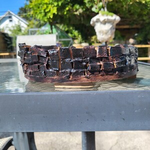 May include: A handmade, low-profile ceramic planter with a dark, textured exterior resembling stacked bricks. The planter has a wide, oval shape and a natural, earthy color palette, suitable for succulents or small plants.