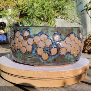 Puede incluir: Un cuenco de cerámica cilíndrico con un patrón de panal texturizado en el exterior. El esmalte muestra una mezcla de tonos marrón tierra, verde bosque profundo y azul vibrante, con las celdas hexagonales en relieve en un acabado bronceado.