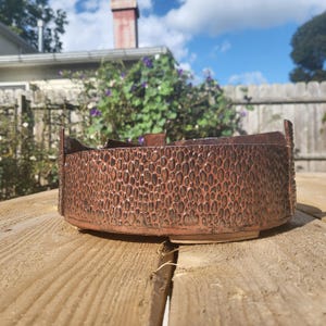 May include: A handcrafted, low-profile ceramic bowl with a textured, reddish-brown exterior. The bowl features a unique pattern of raised, oval-shaped designs and a slightly flared rim. The bowl is sitting on a wooden surface.