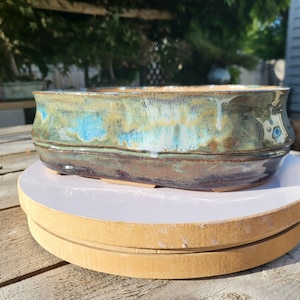 Peut inclure: Un pot de fleurs en céramique de forme ovale avec une finition brillante. Le pot présente un mélange de couleurs bleues, vertes et marron, créant un aspect artistique unique. Le pot est conçu avec un bord légèrement surélevé et repose sur de petits pieds.