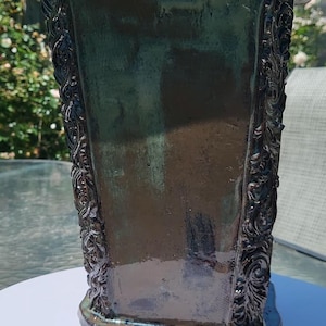 Peut inclure: Un vase en céramique carré et haut avec une finition brillante dans des tons brun foncé et vert profond. Les quatre bords verticaux présentent des motifs en relief complexes qui contrastent avec les surfaces lisses et plates du vase.