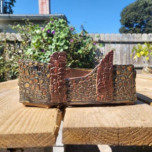 May include: A ceramic planter with a textured, stone-like exterior in shades of brown, black, and gold. The planter has a unique, architectural design with raised sections resembling a fortress. The interior is a darker brown.