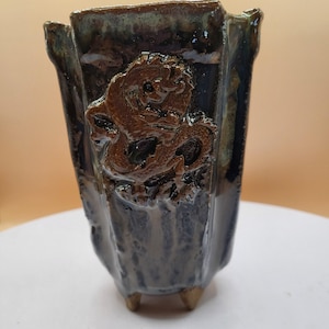 Könnte beinhalten: Handgefertigter Keramik-Pflanztopf mit dunklem, glänzendem Finish. Der Pflanztopf zeigt ein erhabenes, detailliertes Drachendesign in Brauntönen. Das Gefäß hat eine einzigartige, eckige Form und steht auf drei kleinen Füßen.