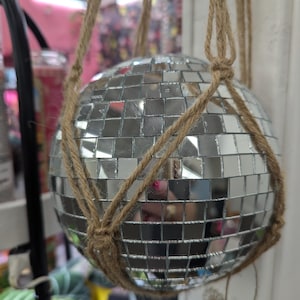May include: A silver disco ball with a mosaic of small mirrored squares hangs from a natural fiber macrame plant hanger. The ball reflects light and surrounding objects. The decorative item is designed for home decor.