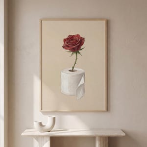 May include: A framed print featuring a red rose in a roll of white toilet paper. The artwork is set against a beige background, with a wooden frame. The image is a still life, with a minimalist aesthetic.