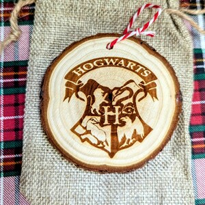May include: A round, wooden ornament with the Hogwarts crest and the word "HOGWARTS" engraved on it. The ornament is made from a natural wood slice and has a red and white string for hanging. It rests on a burlap bag.