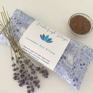 Puede incluir: Una almohada para los ojos azul y blanca con estampado floral y de gatos. La almohada est&aacute; rellena de semillas de lino org&aacute;nico y flores de lavanda. La etiqueta dice "Kendra Veigh Designs" y "Lavender Eye Pillow".