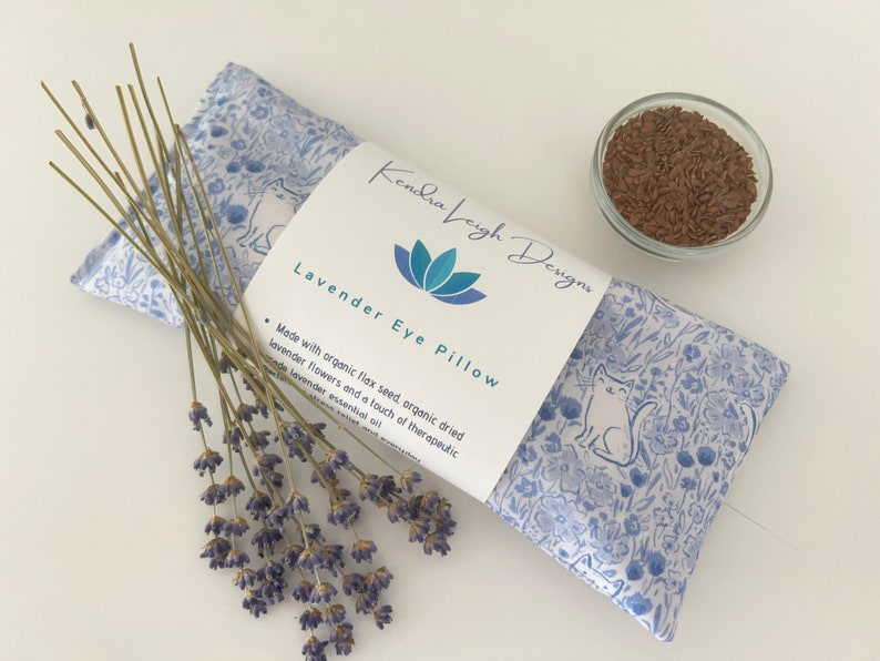 Puede incluir: Una almohada para los ojos azul y blanca con estampado floral y de gatos. La almohada est&aacute; rellena de semillas de lino org&aacute;nico y flores de lavanda. La etiqueta dice "Kendra Veigh Designs" y "Lavender Eye Pillow".
