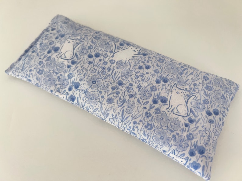 Puede incluir: Una almohada para los ojos azul y blanca con un patr&oacute;n floral y gatos blancos. La almohada es rectangular y tiene una costura en el medio.