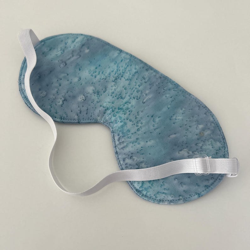 Puede incluir: Una m&aacute;scara para dormir azul con una correa el&aacute;stica blanca. La m&aacute;scara tiene un patr&oacute;n de tie-dye azul claro y blanco.
